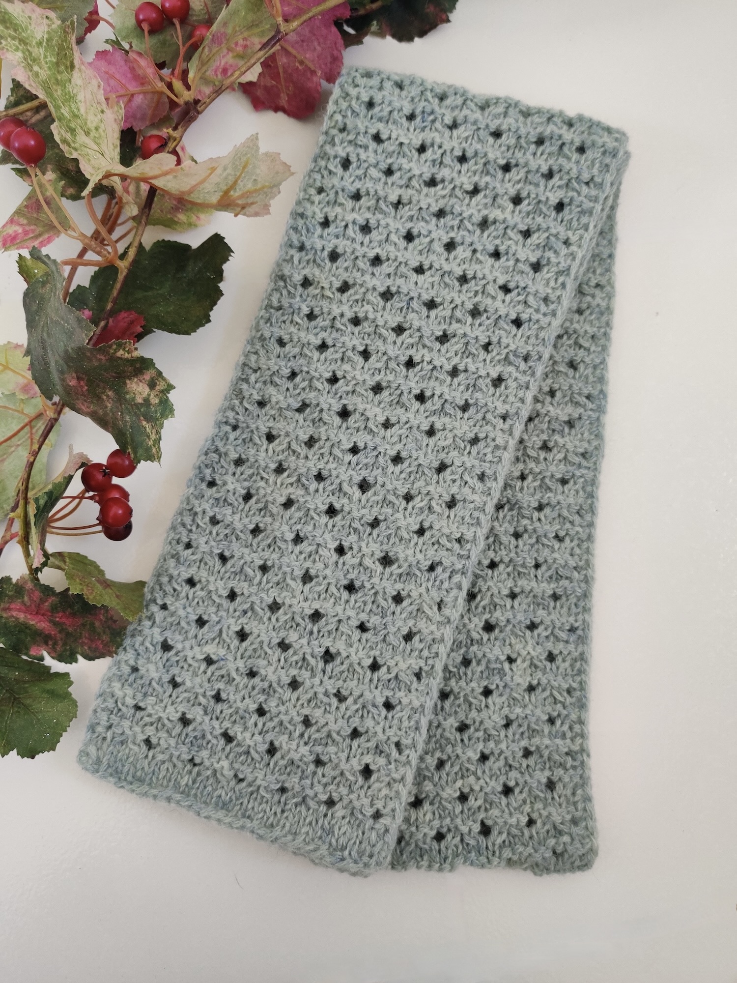 petite écharpe vert d'eau en laine présentée à plat et à côté d'une branche avec des baies